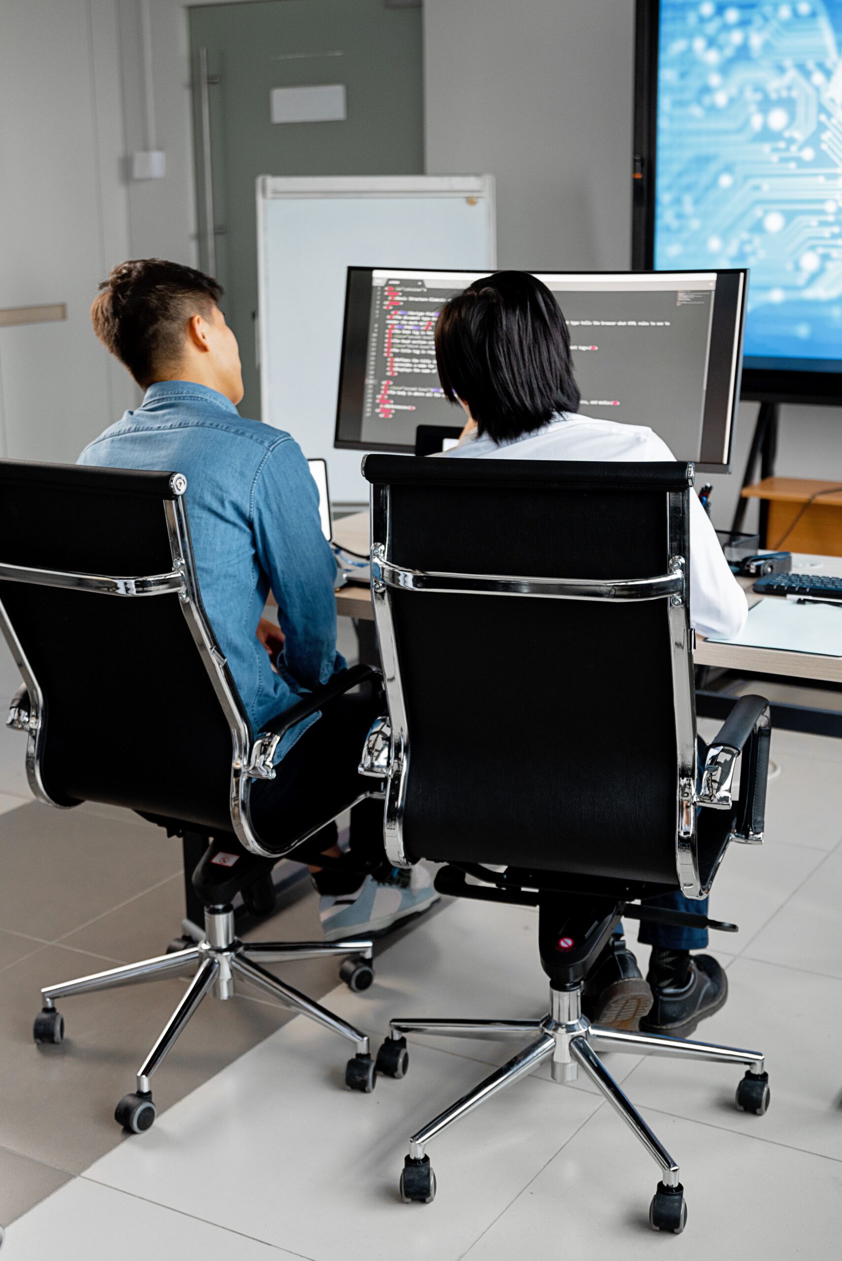 Two professionals collaborating at a desk with dual monitors in an office environment.
